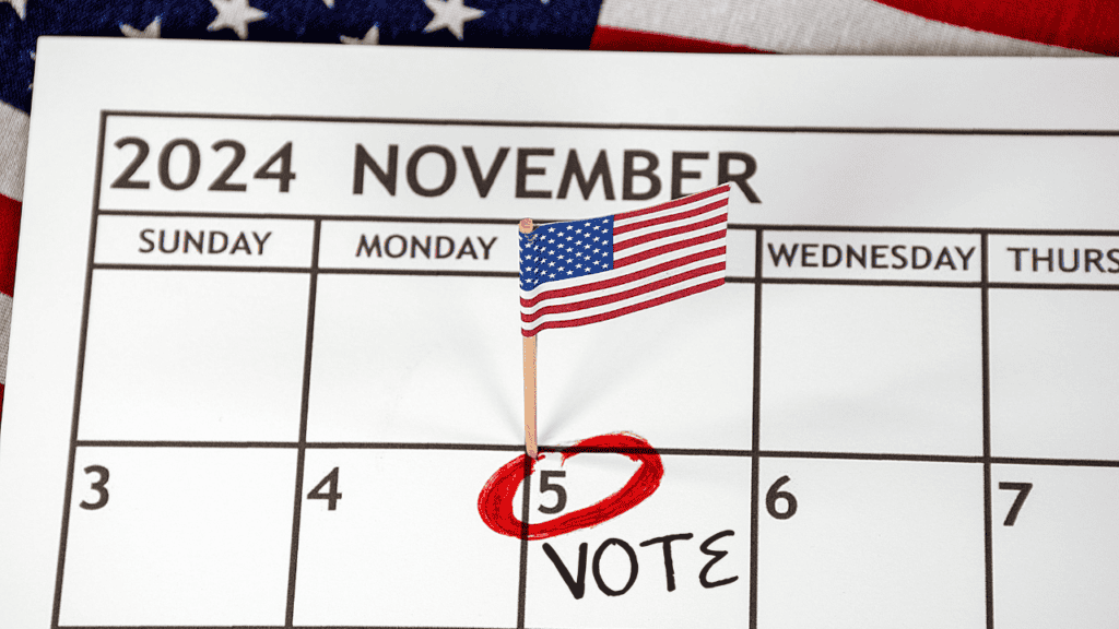 A close-up of a November 2024 calendar page with a small American flag pinned on November 5th, which is circled in red with "VOTE" written underneath. The background shows part of the American flag.