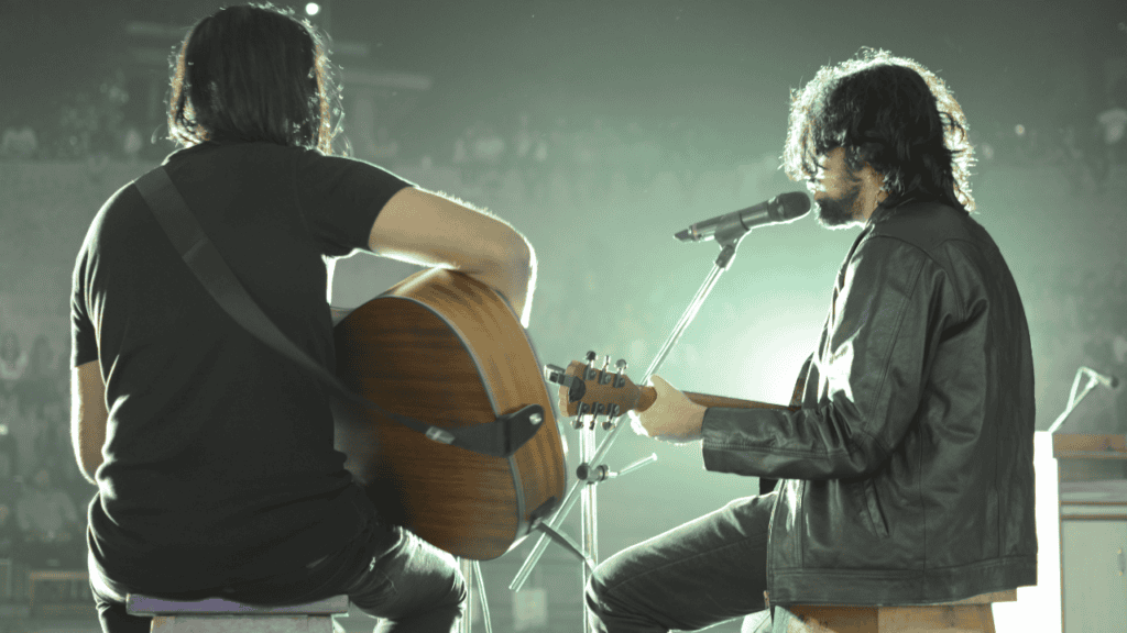 Two musicians performing on stage, one holding an acoustic guitar and the other singing into a microphone. The scene is dimly lit, giving it an intimate atmosphere with a blurred audience in the background.