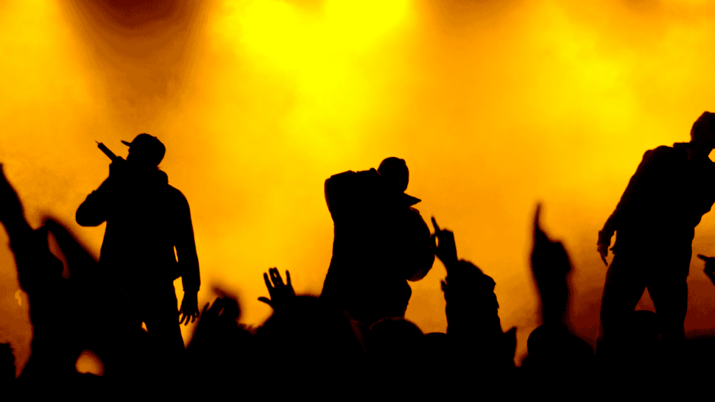 A silhouette of three rappers performing on stage against a vibrant orange background with an audience reaching out towards them, capturing the energy of a live concert.