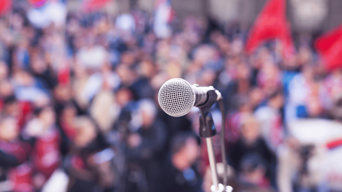 A microphone on a stand in focus, with a blurred crowd in the background, suggesting a rally, protest, or public speech setting.