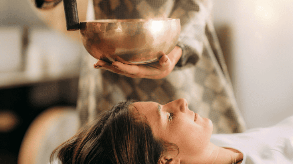 A woman lying down during a sound therapy session, with a sound bowl being played above her.