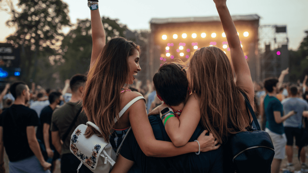 Three friends embracing at a music festival with the stage glowing in the background.