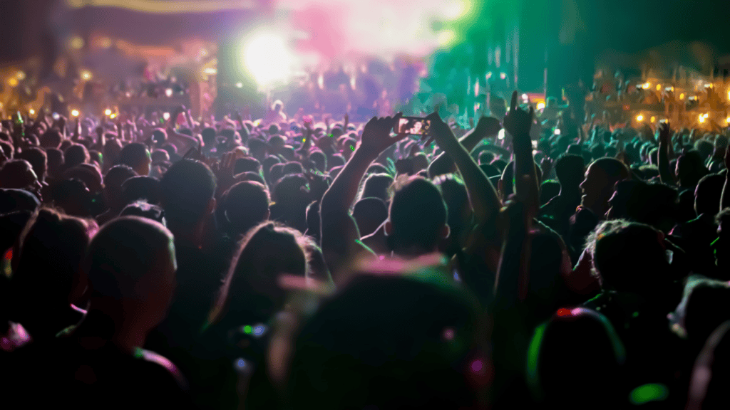 Nighttime crowd at a live concert, illuminated by vibrant stage lights and a phone capturing the moment.