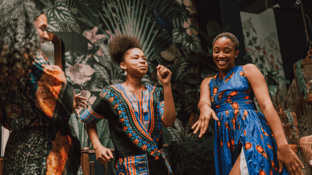Two women smiling and dancing in colorful cultural attire indoors.