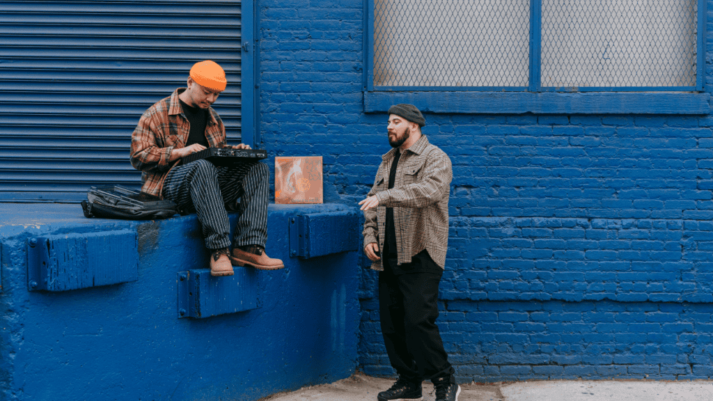 Two men on an urban blue backdrop creating music with a laptop and beat pad.