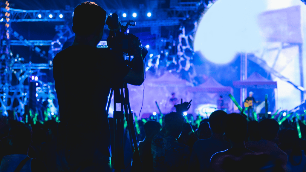 A silhouette of a cameraman recording a live concert with a vibrant blue stage in the background.