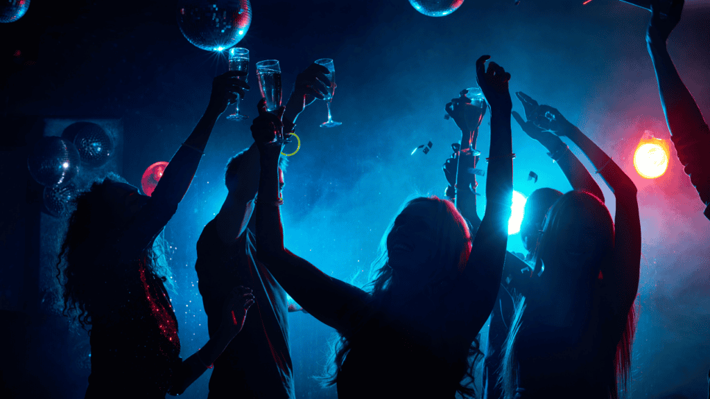Silhouetted partygoers raising glasses in a lively nightclub with disco balls overhead.
