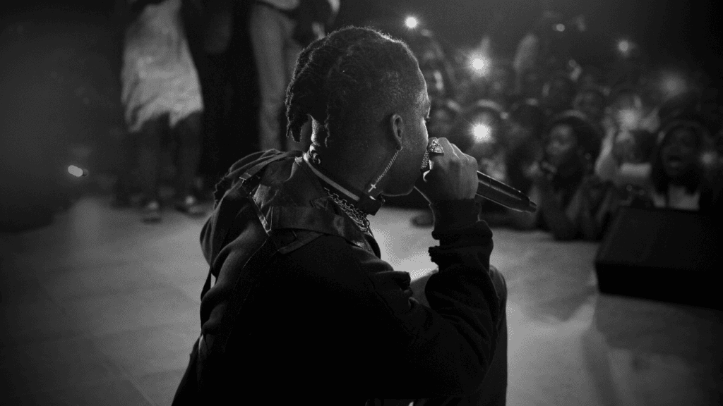 A hip-hop artist kneeling on stage, performing to an enthusiastic crowd in black and white.