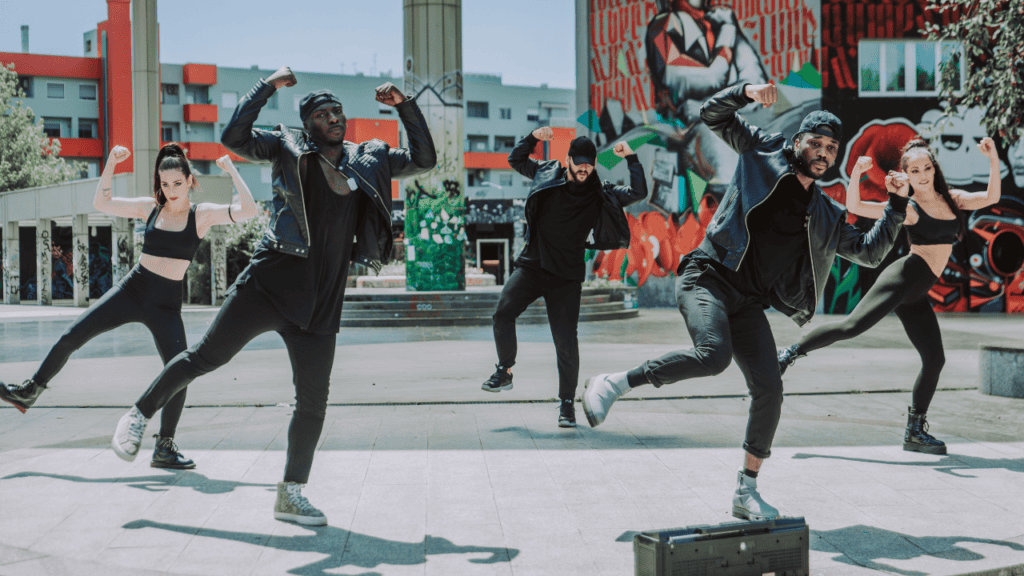 A group of dancers performing a choreographed routine in an urban plaza with colorful graffiti.