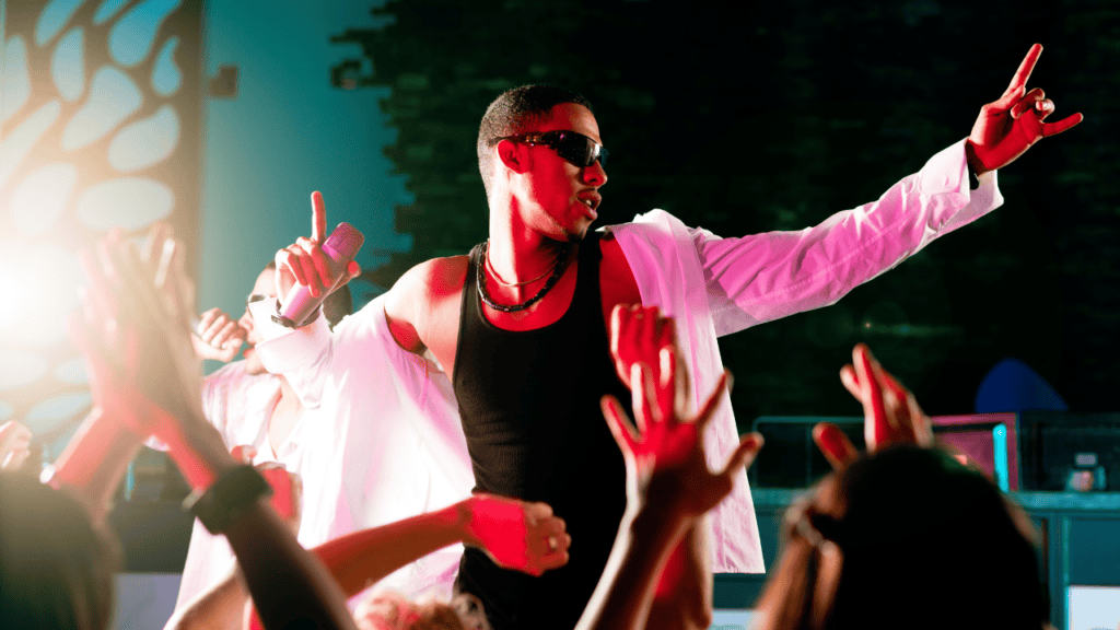 A performer in a white jacket raising his arms on stage, hyping up a dancing crowd.
